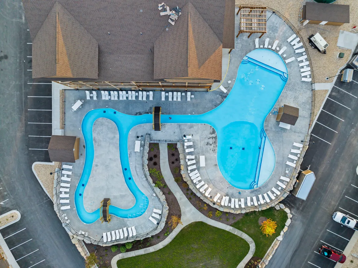 An exterior scene showing two uniquely shaped swimming pools surrounded by lush landscaping and a building adjacent to the water. The pools are active with social gatherings and sunbathers, evoking the playful, relaxed resort vibe.