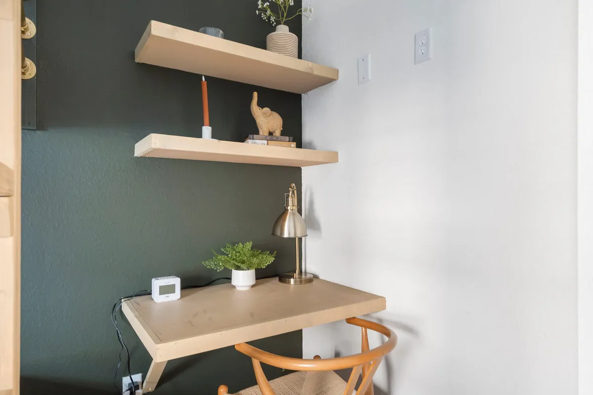 A home office setup featuring a wooden desk, shelves with books and decor, a lamp, and a potted plant. The space is calm, organized, and perfect for remote work or reading in peace.