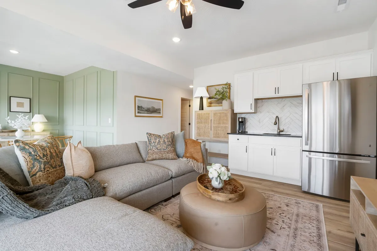 A welcoming entertainment space with a modern sectional sofa, round coffee table, decorative pillows, and a kitchenette area in the background. The layout is ideal for grabbing snacks, socializing, or enjoying a casual meal.