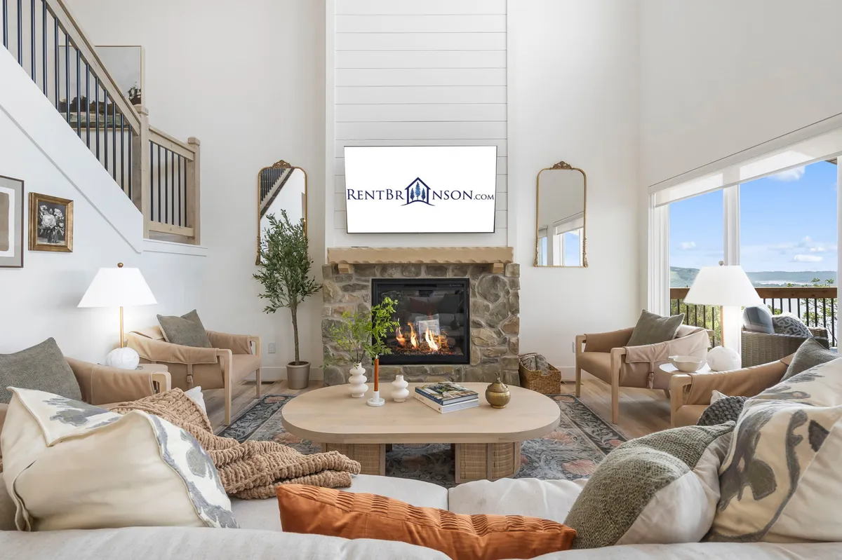 A cozy living room featuring an elegant fireplace, a comfortable sofa, a coffee table, and large windows that allow sunlight to flood the area. The setup is intimate and oriented for both relaxation and social gatherings, with the windows likely offering glimpses of scenic outdoor views.