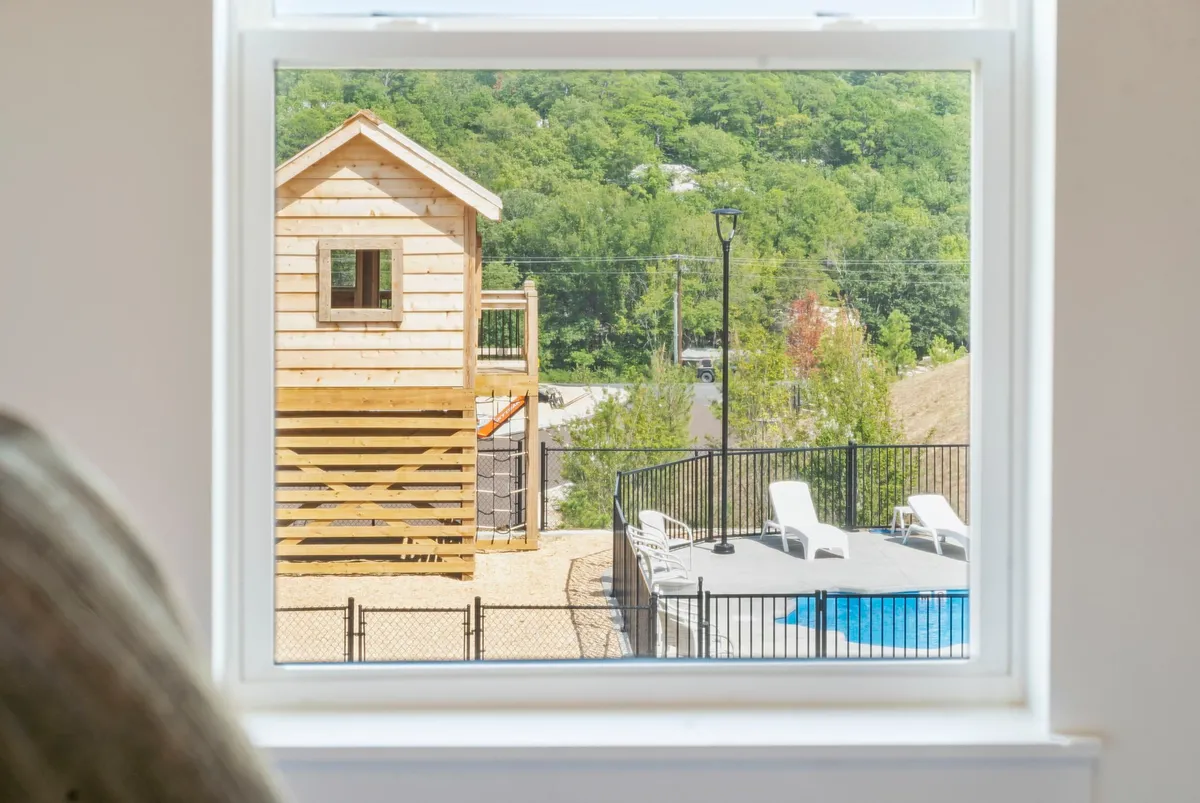 This vibrant exterior scene features a wooden playhouse, a sparkling swimming pool surrounded by sun loungers, and lush greenery. Children can be seen playing near the playhouse, while adults relax poolside, creating an atmosphere of family fun and outdoor togetherness.