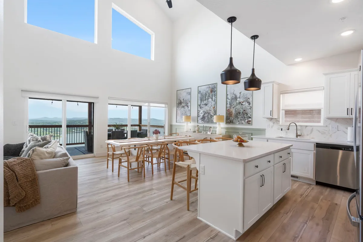 An expansive, open interior with a large dining table, kitchen island, and dramatic floor-to-ceiling windows. Artwork adorns the walls, and the space is styled for both comfort and elegance, with ample room for group gatherings or family meals against a backdrop of scenic views.