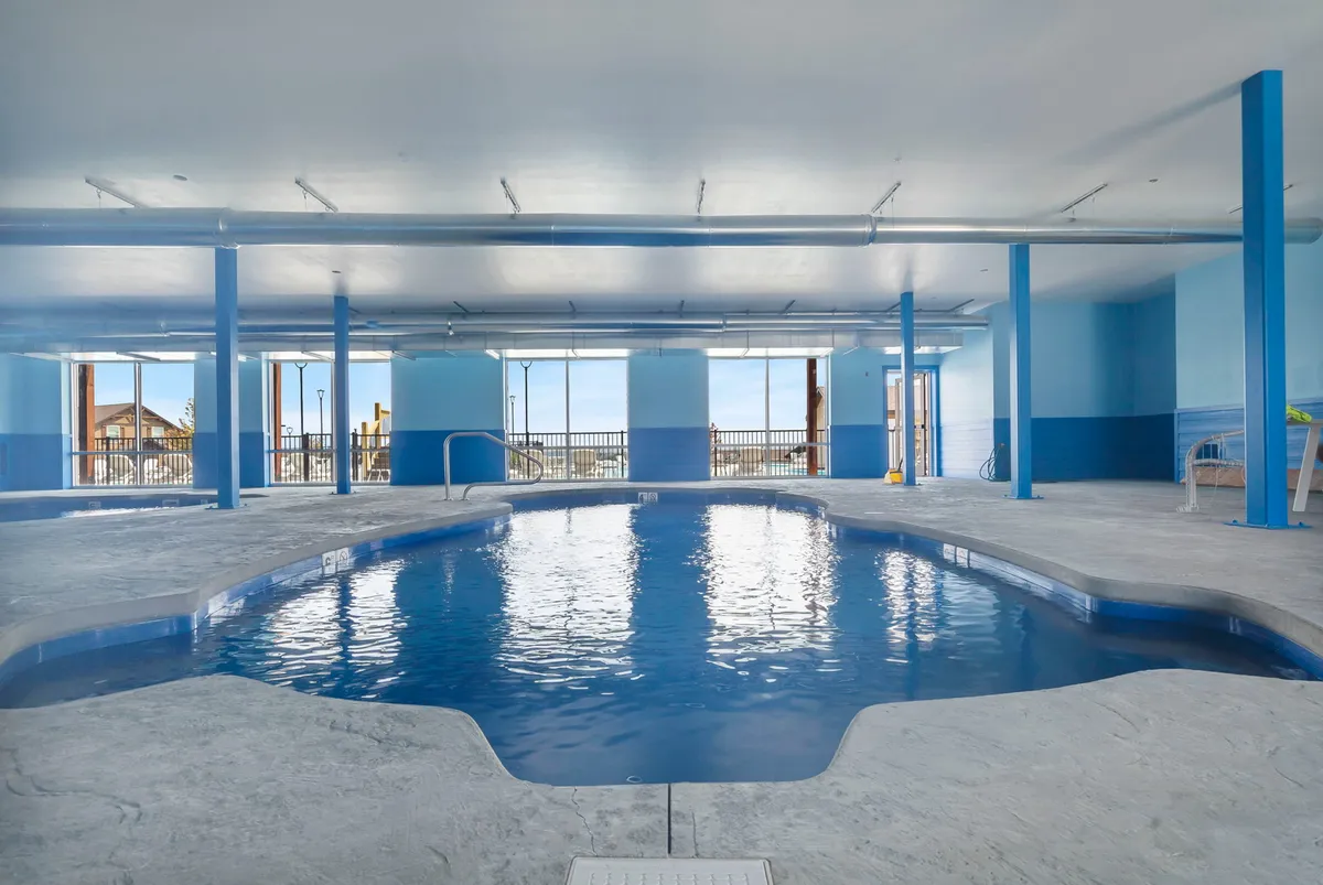 An inviting indoor swimming pool surrounded by large windows with blue walls, creating a tranquil and light-filled space for swimming, relaxing, or socializing. The setting suggests opportunities for wellness and rejuvenation, rain or shine.