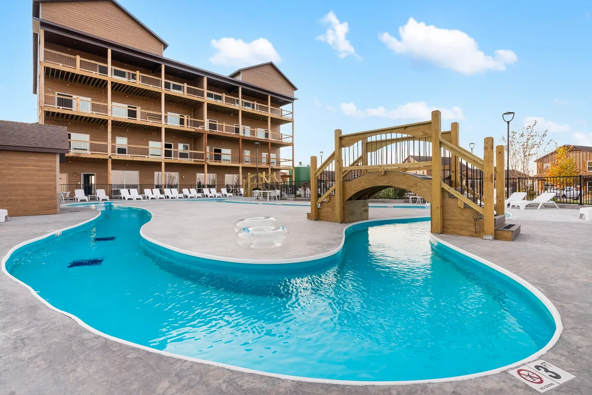 The exterior of the property is showcased with a luxurious swimming pool, a wooden bridge, and comfortable lounge chairs. The backdrop includes the main building, and the scene is set at sunset, enhancing the sense of relaxation and gathering.