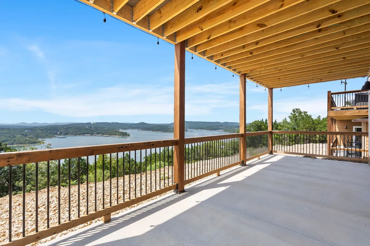 A spacious wooden deck stretches toward an expansive lake view, with open-air seating and space for outdoor dining or sunbathing. The pergola structure overhead adds architectural interest and shade, while the tranquil water and distant hills create a sense of serenity and escape.
